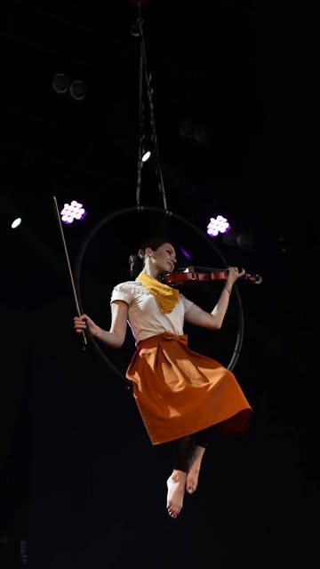 A violinist plays while seated in an aerial hoop suspended above a dark stage, lit by scattered purple lights.
