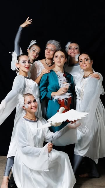 Eight performers grouped against black: seven in white, one at the centre in teal holding a violin and a white feather, with a white dove held out in front.