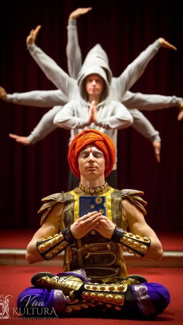 A performer in a saffron turban and gold armour sits cross-legged in meditation; behind, dancers in white extend their arms to form a many-armed silhouette against a deep red backdrop.